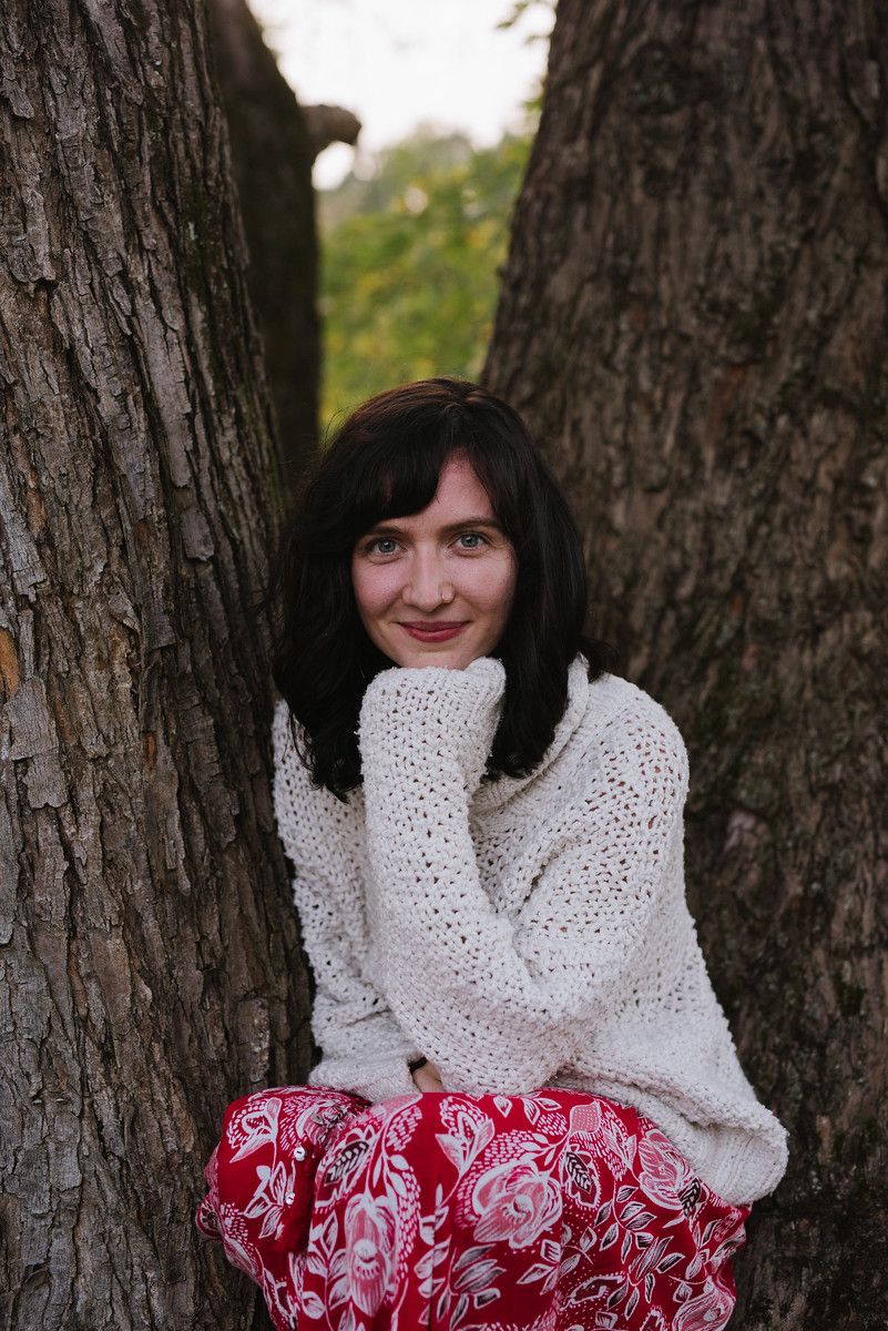 Nuala Schoen in a white knit sweater and red floral with a hand on her chin smiling in front of a tree