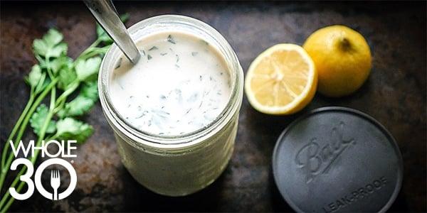 A jar of Whole30 Lemon Tahini dressing is shot from above, styled next to fresh herbs and a fresh lemon.