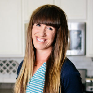 A headshot of Autumn Michaelis smiling, wearing a light blue shirt with a navy blue cardigan. In the background of the shot is a white kitchen