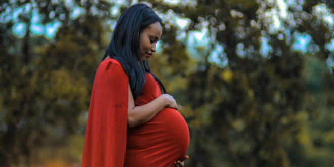 A pregnant Black woman in a red dress holds her belly and looks down. She is standing in front of some trees and a blue sky.