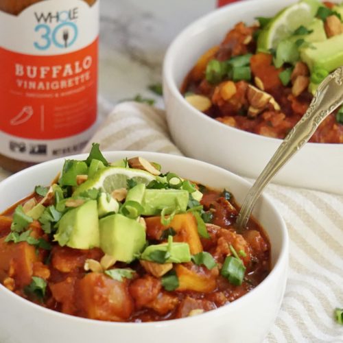 Two bowl of buffalo chicken chili, toped with avocado and cilantro. Behind the two bowls are two bottles of the Whole30 Buffalo Vinaigrette dressing and marinade.