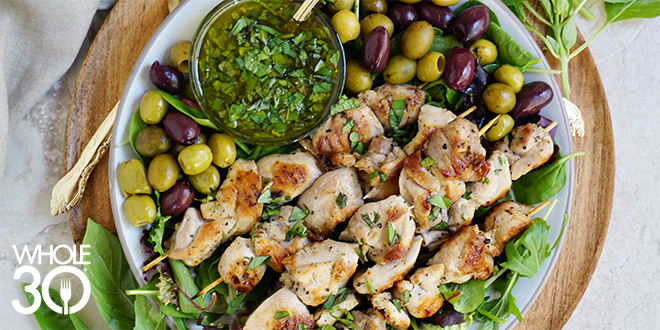 Chicken skewers on a bed of herbs and olives. Next the skewers is a small dish of basil cilantro mint sauce.