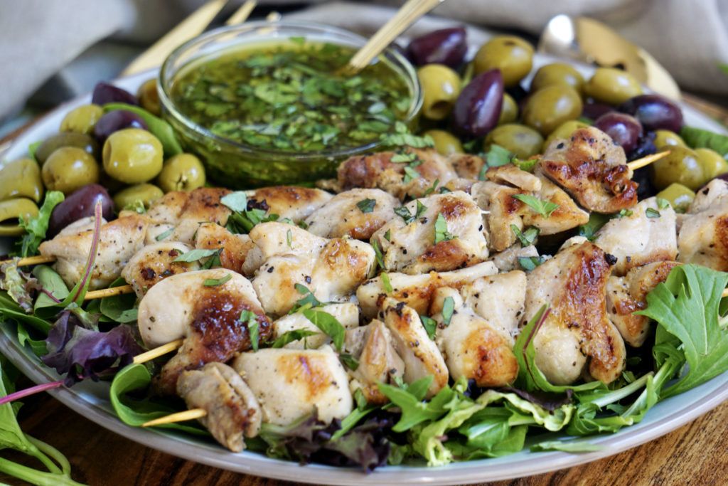Chicken skewers on top a bed of fresh greens and olives. Next to the skewers is a basil, cilantro, mint sauce is a small dish. 