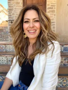Jackie Jorge in a black top, white sweater, and jeans sitting on stairs and smiling.