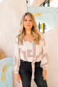 Alyssa smiling and standing with hands by her side, wearing a cream patterned shirt and black jeans.