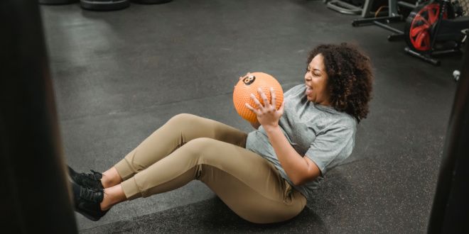 A woman smiles as she holds boat pose with a medicine ball in her hands
