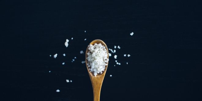 A wooden spoon full of big flakes of salt rests on a black background with some salt flakes scattered about.