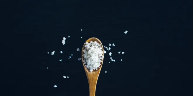 A wooden spoon full of big flakes of salt rests on a black background with some salt flakes scattered about.