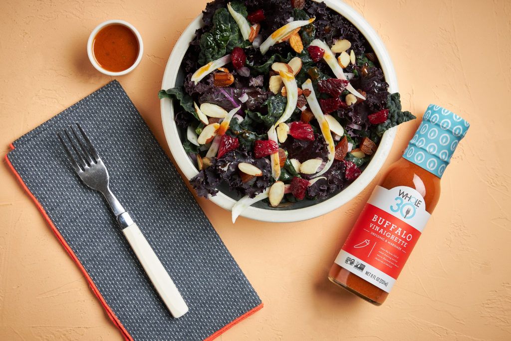 A bowl of Citrus Kale Salad next to a bottle of Made by Whole30 Buffalo Vinaigrette.