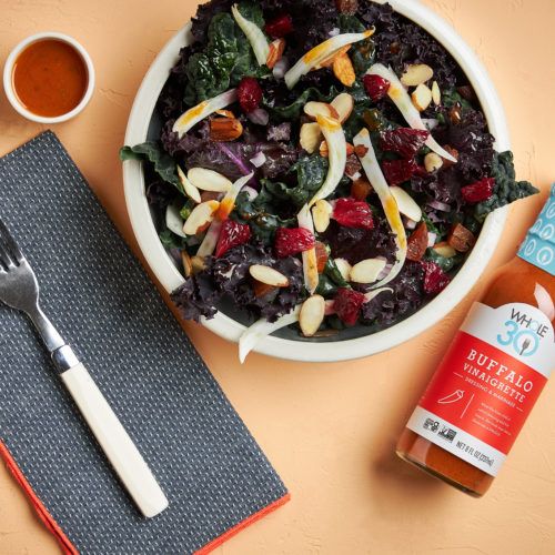 A bowl of Citrus Kale Salad next to a bottle of Made by Whole30 Buffalo Vinaigrette.