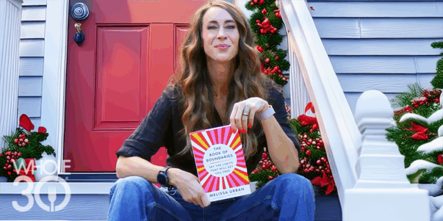 A smiling woman with long hair, sitting on a holiday-themed front porch with a red front door. She is holding The Book of Boundaries.