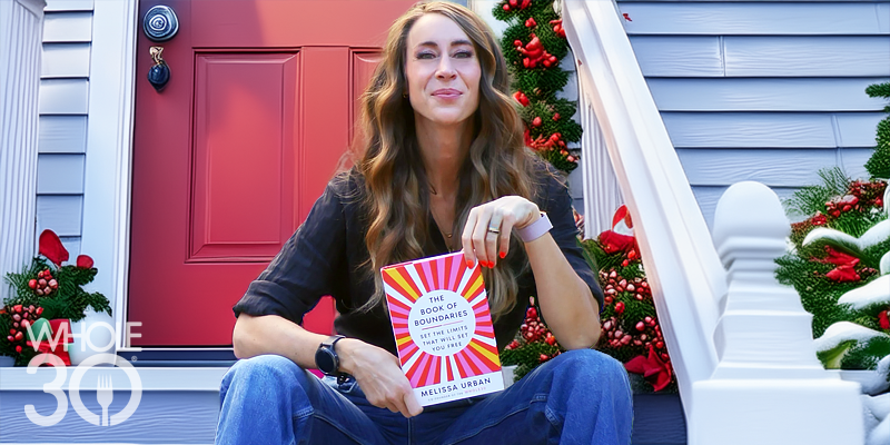 A smiling woman with long hair, sitting on a holiday-themed front porch with a red front door. She is holding The Book of Boundaries.