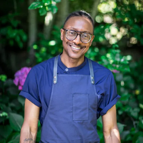 Gregory Gourdet wearing a blue shirt and a navy apron, smiling at the camera.