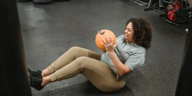 A woman smiles as she holds boat pose with a medicine ball in her hands