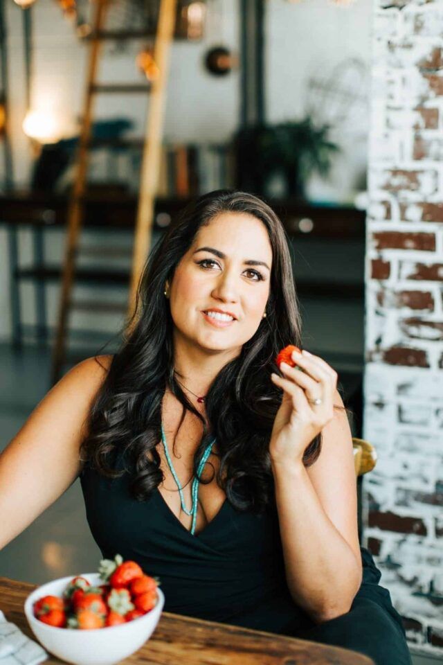 Cristina Curp sitting at a table with a bowl of strawberries. She is holding a strawberry in one of her hands and is smiling at the camera.