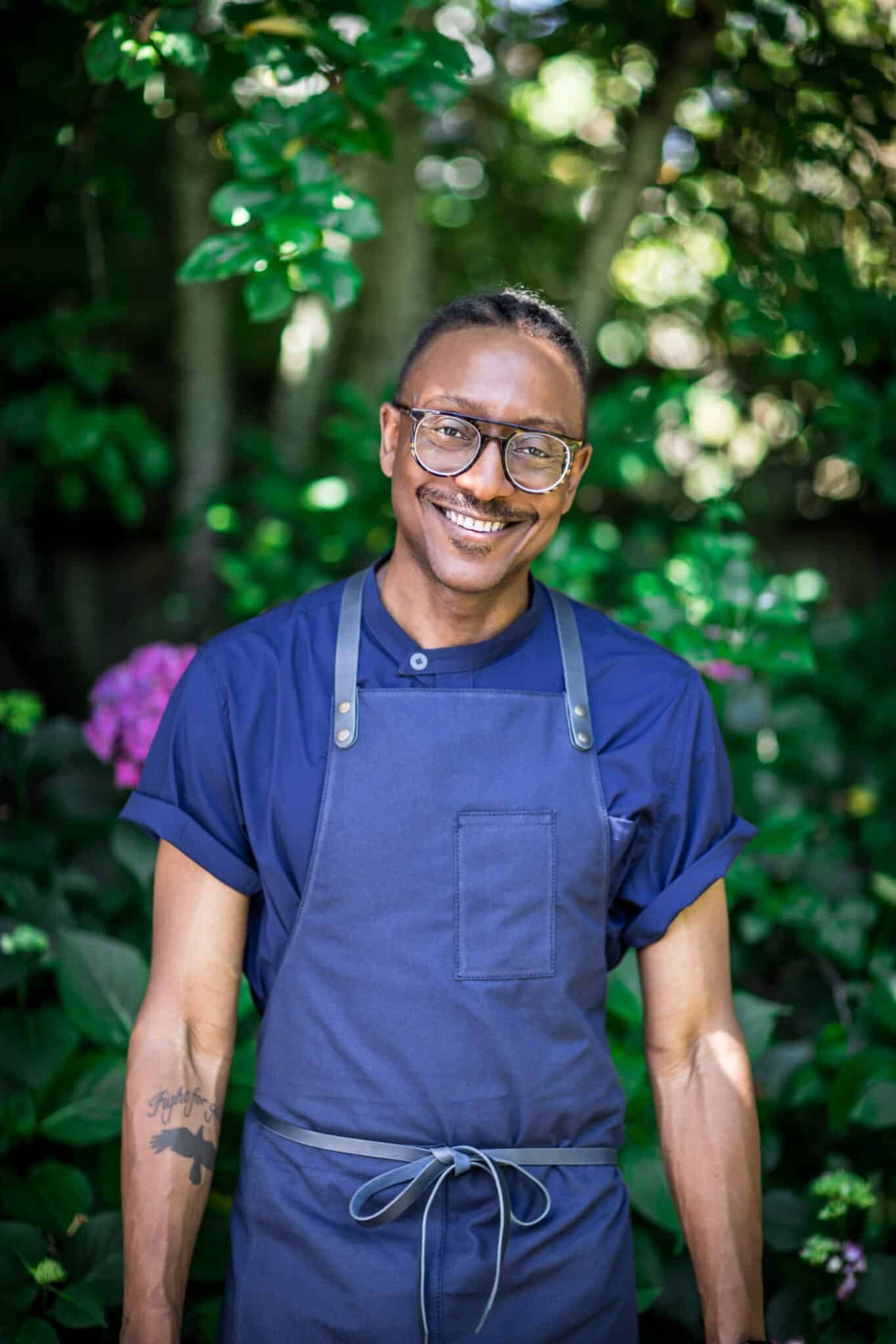 Gregory Gourdet wearing a blue shirt and a navy apron, smiling at the camera.
