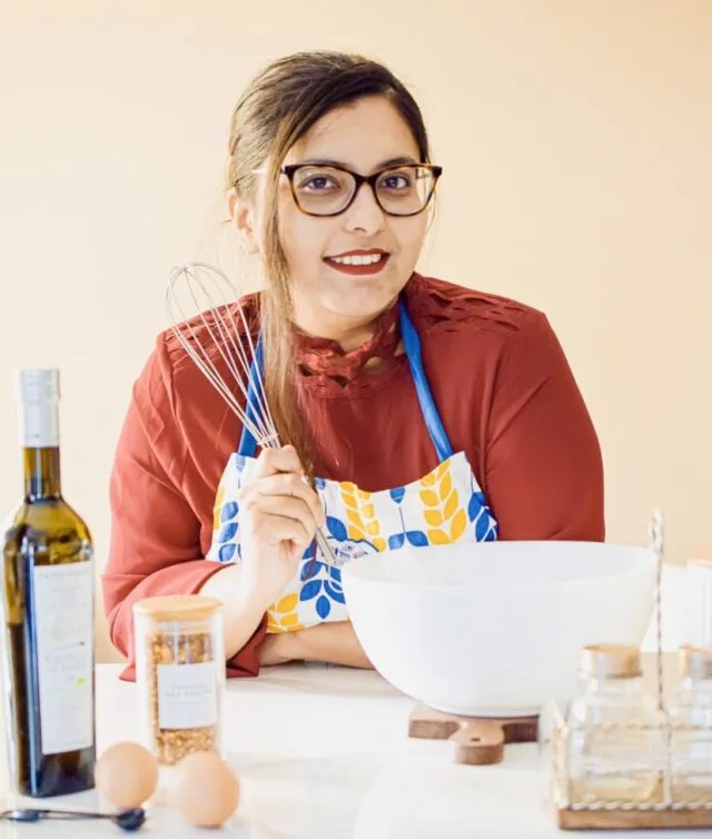 Ruda Khan of My Gourmet, in an orange red stop with an apron smiling at the camera with a whisk in her hand.