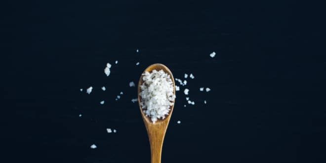 A wooden spoon full of big flakes of salt rests on a black background with some salt flakes scattered about.