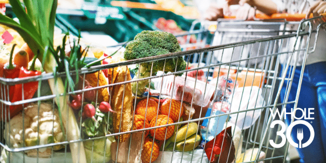 A cart full of Whole30 compatible produce and essentials is being pushed through a grocery store by a couple. The cart is prominent in the foreground and you can only see the couple out of focus in the background.