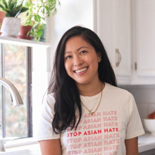 Jean Choi wearing a white shirt that reads "Stop Asian Hate" in red lettering, in a kitchen smiling at the camera.