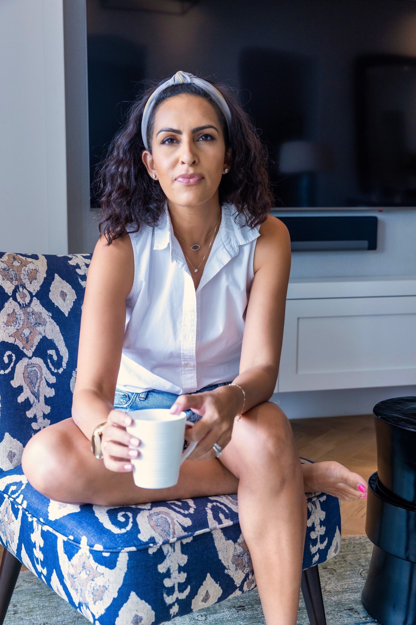 Ryani Rupani wearing a white shirt, sitting in a chair with a coffee mug, smiling at the camera.