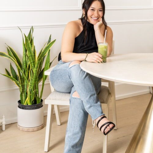 Jessica Sussman , a recipe and content creator, wearing blue jeans and a black tank top. She is sitting down at a tan table, and is smiling at the camera.