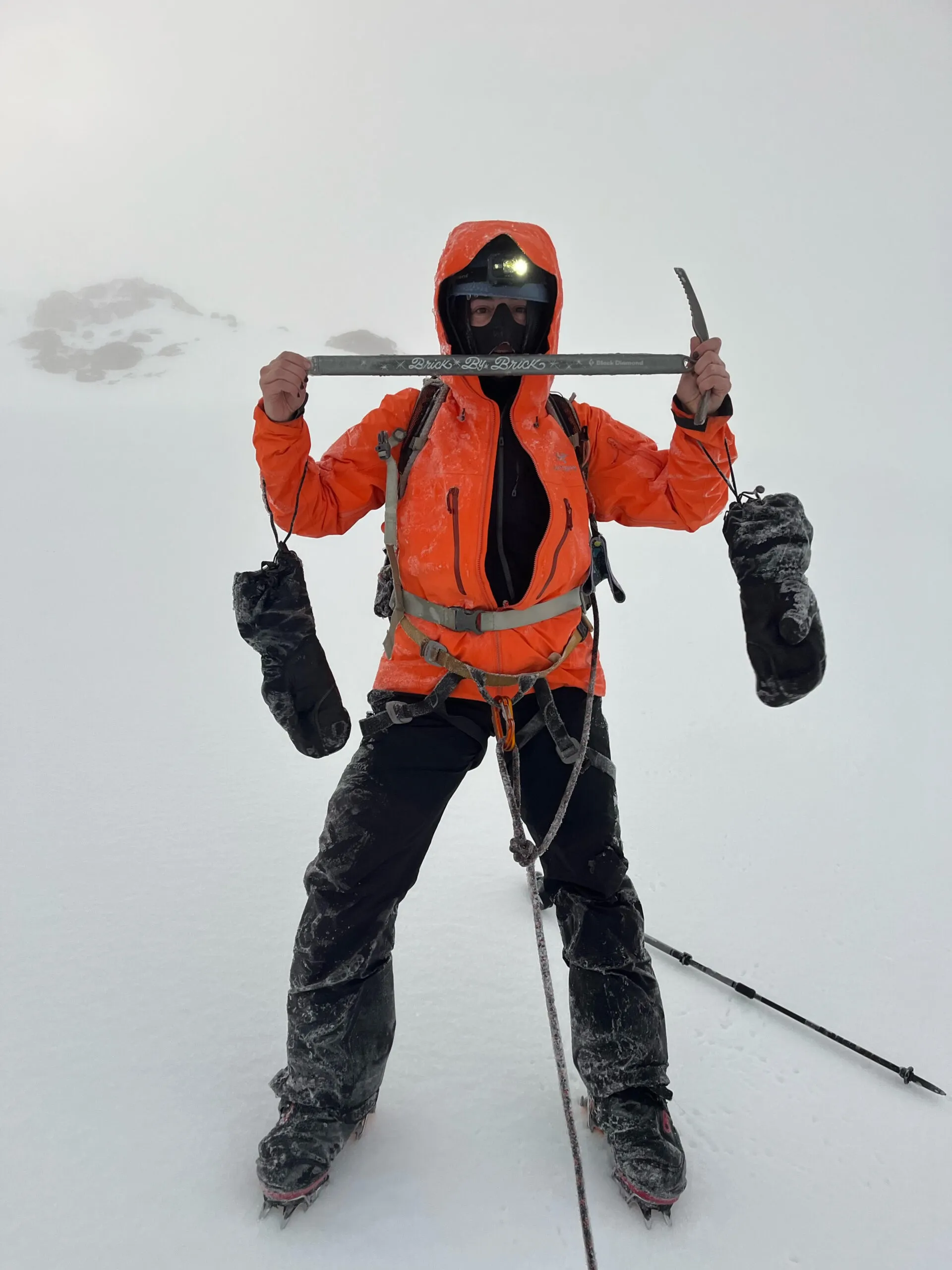 Sarah in an orange winter jacket with black pants holding up an ice axe.