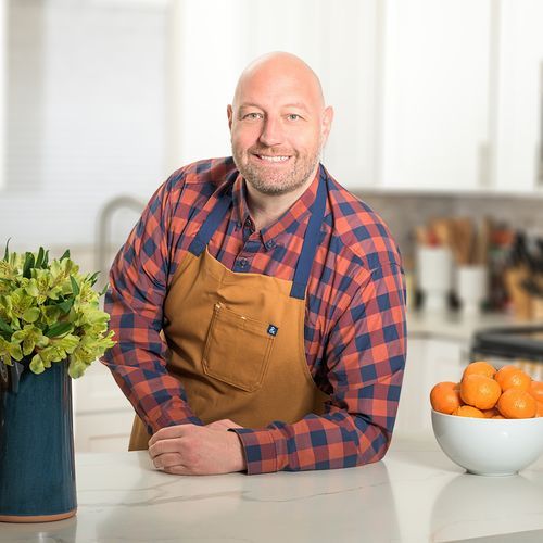 Reed Dunn of Pesto and Potatoes, wearing a red and blue flannel underneath a brown apron, leaning on a kitchen counter and smiling at the camera.
