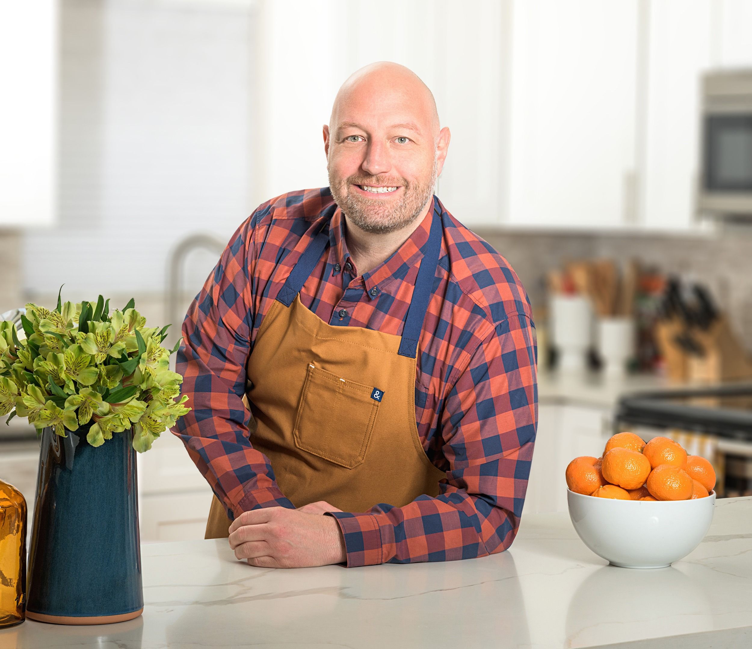 Reed Dunn of Pesto and Potatoes, wearing a red and blue flannel underneath a brown apron, leaning on a kitchen counter and smiling at the camera.