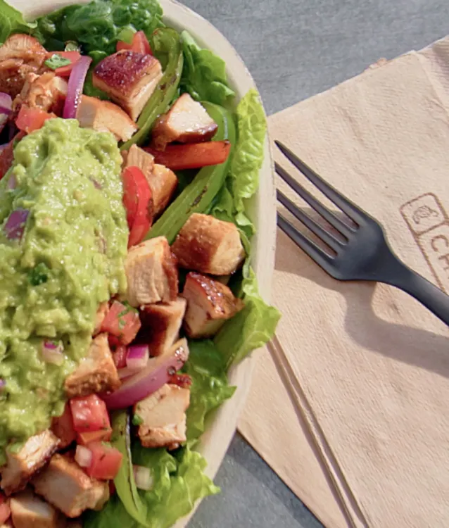 A well lit chicken adobo bowl with a heap of guacamole sitting atop a Chipotle napkin with a black fork slightly to the right. A reddish tan leather spiral notebook sits to the top left of the bowl, falling mostly out of frame.