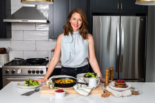 Dr.Casey Means in a light blue tank top and black pants, is standing at a white marble kitchen counter with food on the counter. She is smiling at the camera.