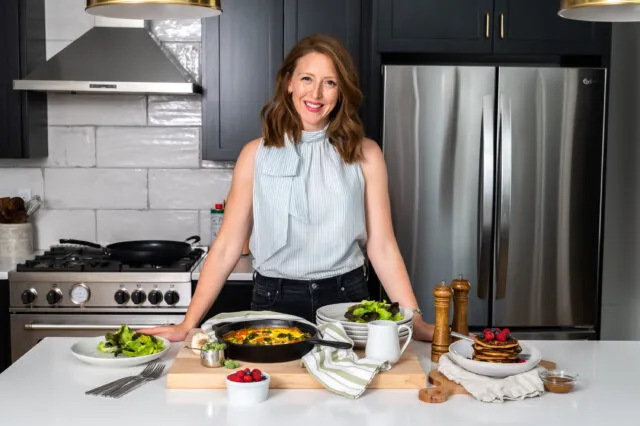 Dr.Casey Means in a light blue tank top and black pants, is standing at a white marble kitchen counter with food on the counter. She is smiling at the camera.