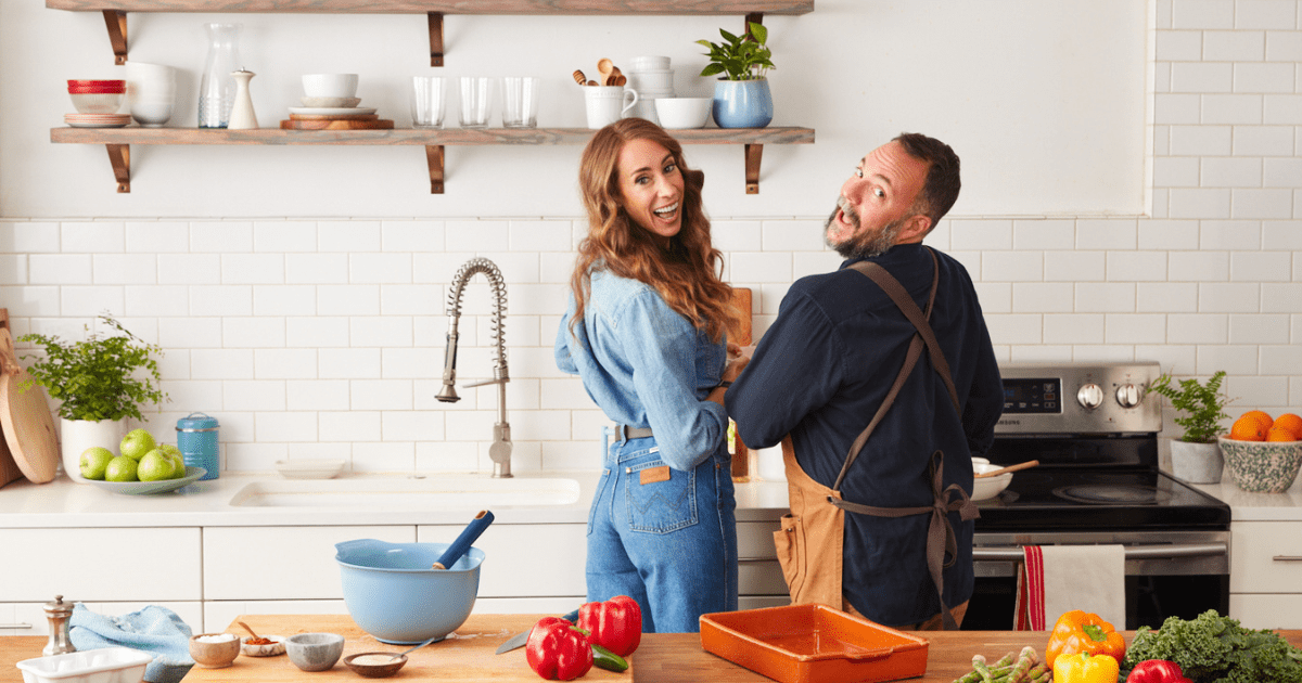 A behind the scenes photo of The New Whole30 showing Melissa Urban and Barrett Washburne next to each other in a kitchen, their backs to the camera, turning their faces towards the camera and smiling