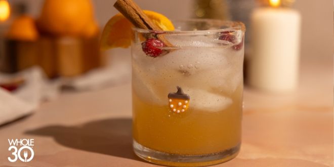 Apple Cinnamon Mockarita on a mabrle backround with a cinnamon stick, orange, and cranberry garnish. The glass has an acorn on it, and a candle is lit in the background.