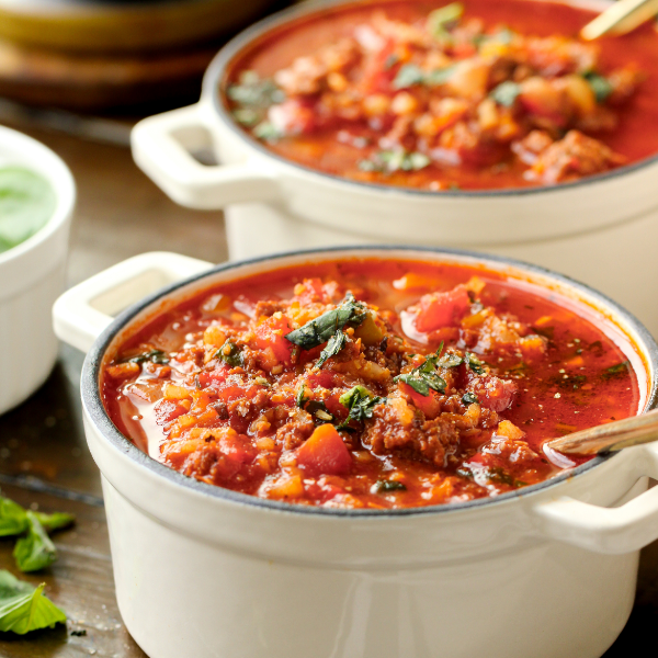 Hearty red stuffed pepper soup in two white ramekins, topped with fresh green herbs on wooden background