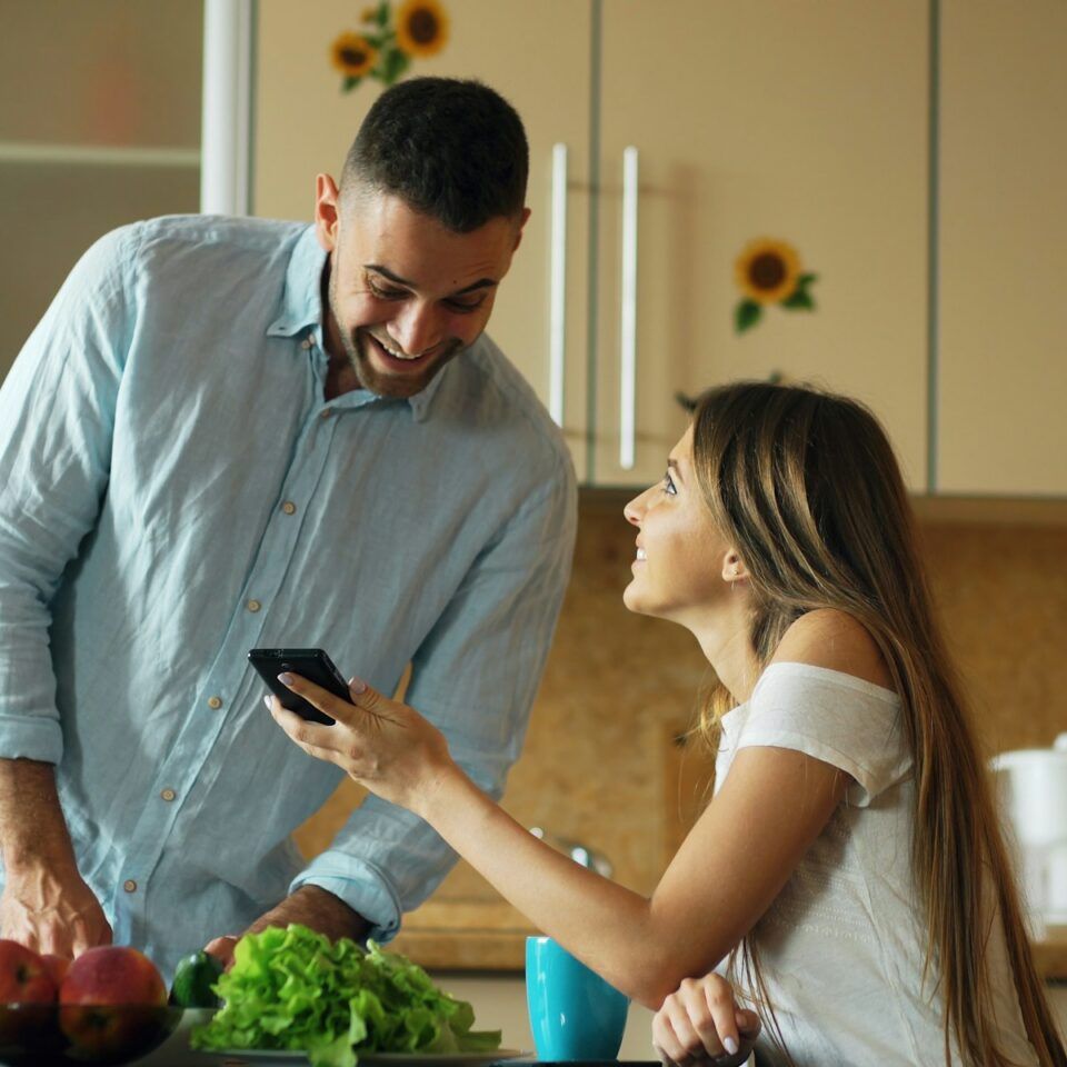 Couple smiling together over a phone in a bright kitchen, with lettuce and apples on the counter
